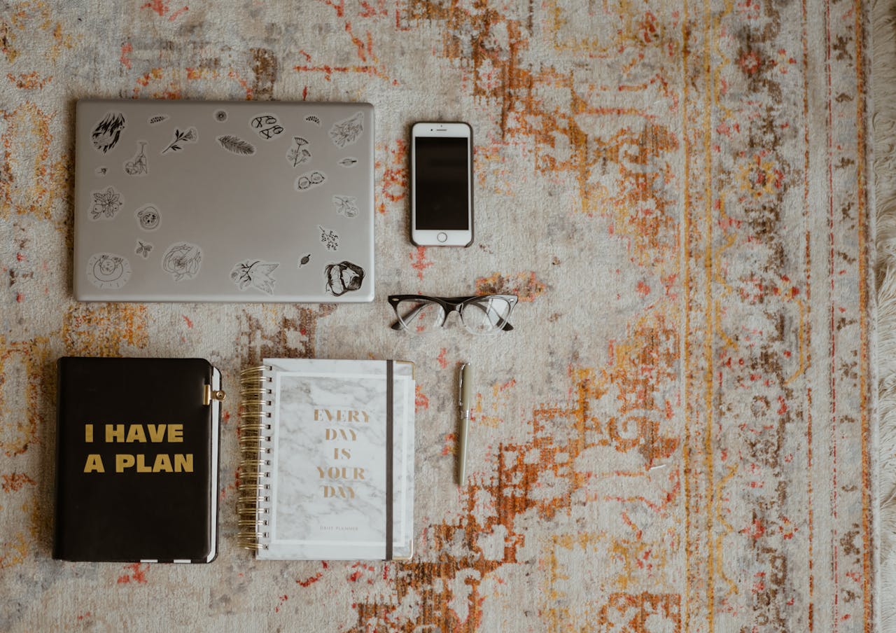 who-we-are-img Flat lay of workspace items including laptop, phone, planners, and glasses on a patterned rug.